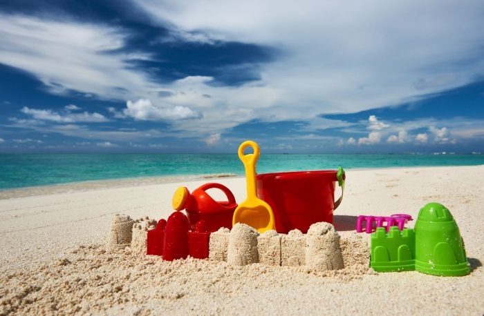 Colorful layered paper cut-out collage of beach toys and sandcastles on turquoise shoreline under a blue sky, playful and bright mood